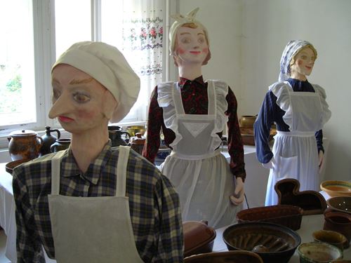 Hölzerne Puppen in Lebensgröße sind in Damenhemden und Schürzen gekleidet. Um sie herum befinden sich zahlreiche alte Schalen, Töpfe und Kuchenformen.