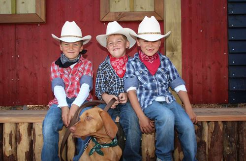 Drei Jungen im Cowboy-Outfit, die auf einer Holzbank sitzen. Einer hält die Leine eines braunen Weimaraners fest.