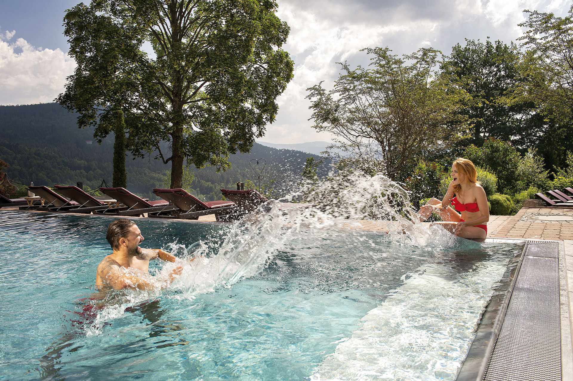 Ein Paar bespritzt sich in einem Pool mit Wasser. Im Hintergrund ist eine Panoramaaussicht des Bayerischen Waldes.
